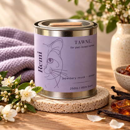 A tawne candle tin with muted lavender mauve label showing image of a cat. the name remi is labeled. sat on a stone coaster with decorative flowers and amber stone around.