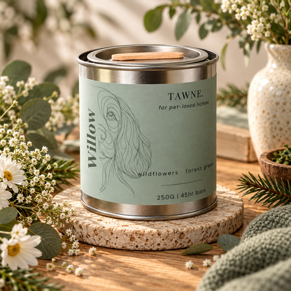 A metal tin candle labeled 'TAWNE' by Willow, styled on a stone coaster with flowers, greenery, and a knit blanket in a warm indoor setting.