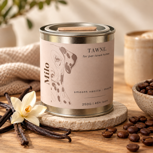 A metal tin candle labeled TAWNE for pet-loved homes, Milo, in a styled tabletop setting with vanilla beans, a flower, coffee beans, and a woven cloth.