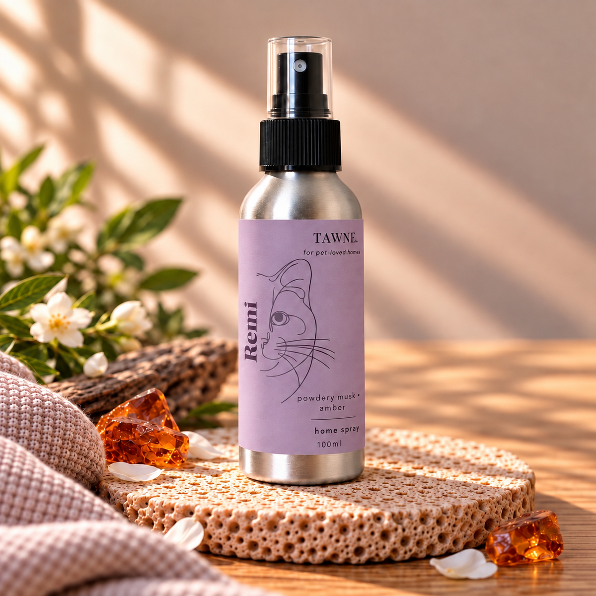 A 100ml aluminum home spray bottle with a lavender label by Tawne, styled on a textured stone coaster with amber crystals and white blossoms on a wooden surface.