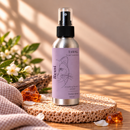 A 100ml aluminum home spray bottle with a lavender label by Tawne, styled on a textured stone coaster with amber crystals and white blossoms on a wooden surface.