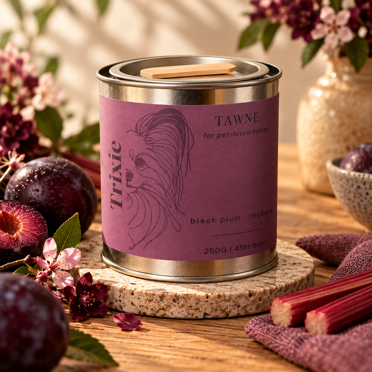 A tin Trixie Tawne pet safe candle, labeled 'black plum - rhubarb', styled on a wooden table with plums, rhubarb stalks, and flowers.