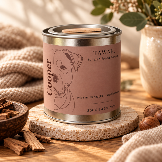 A metal tin candle labeled 'Cooper' and 'TAWNE' sits on a stone coaster on a wooden table, surrounded by cozy props including a knit blanket, cinnamon sticks, nuts, and dried flowers.
