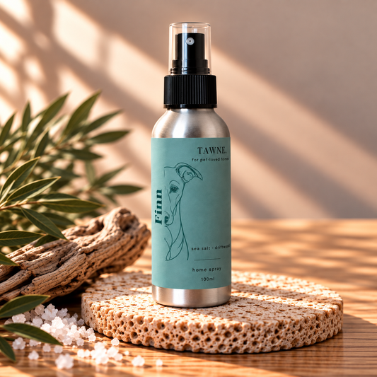 A silver metal home spray bottle with a black spray top and a light blue-green label reading TAWNE and Finn, styled on a woven mat with bath salts, driftwood, and greenery in warm sunlight.