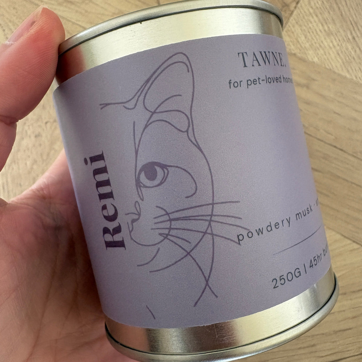 A hand holding a silver metal tin Remi Tawne candle, showing the lavender label with a minimalist cat line drawing, against a wooden floor background.