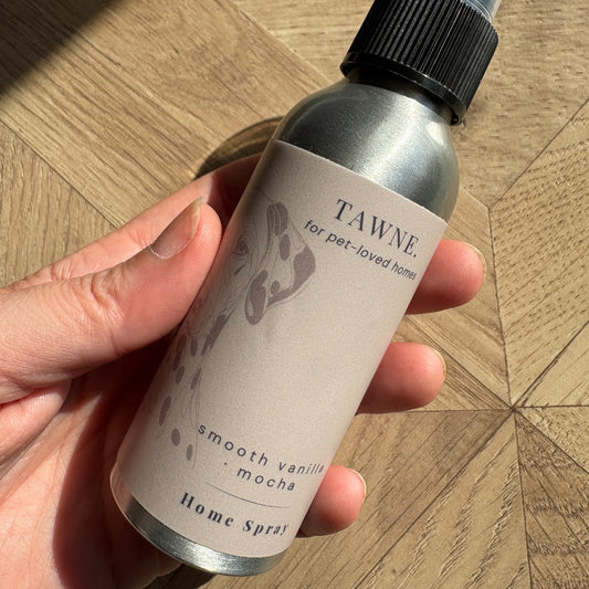 A hand holding a silver metal bottle of Tawne Home Spray, featuring a black spray nozzle and a beige label with vanilla and mocha scent details, set against a wooden parquet floor.