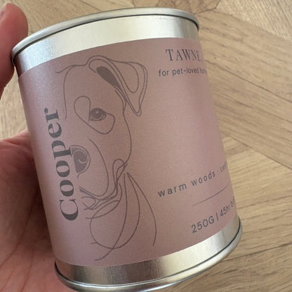 A hand holding a cylindrical metal tin ofA  Cooper Tawne candle
 on a light wood floor background.