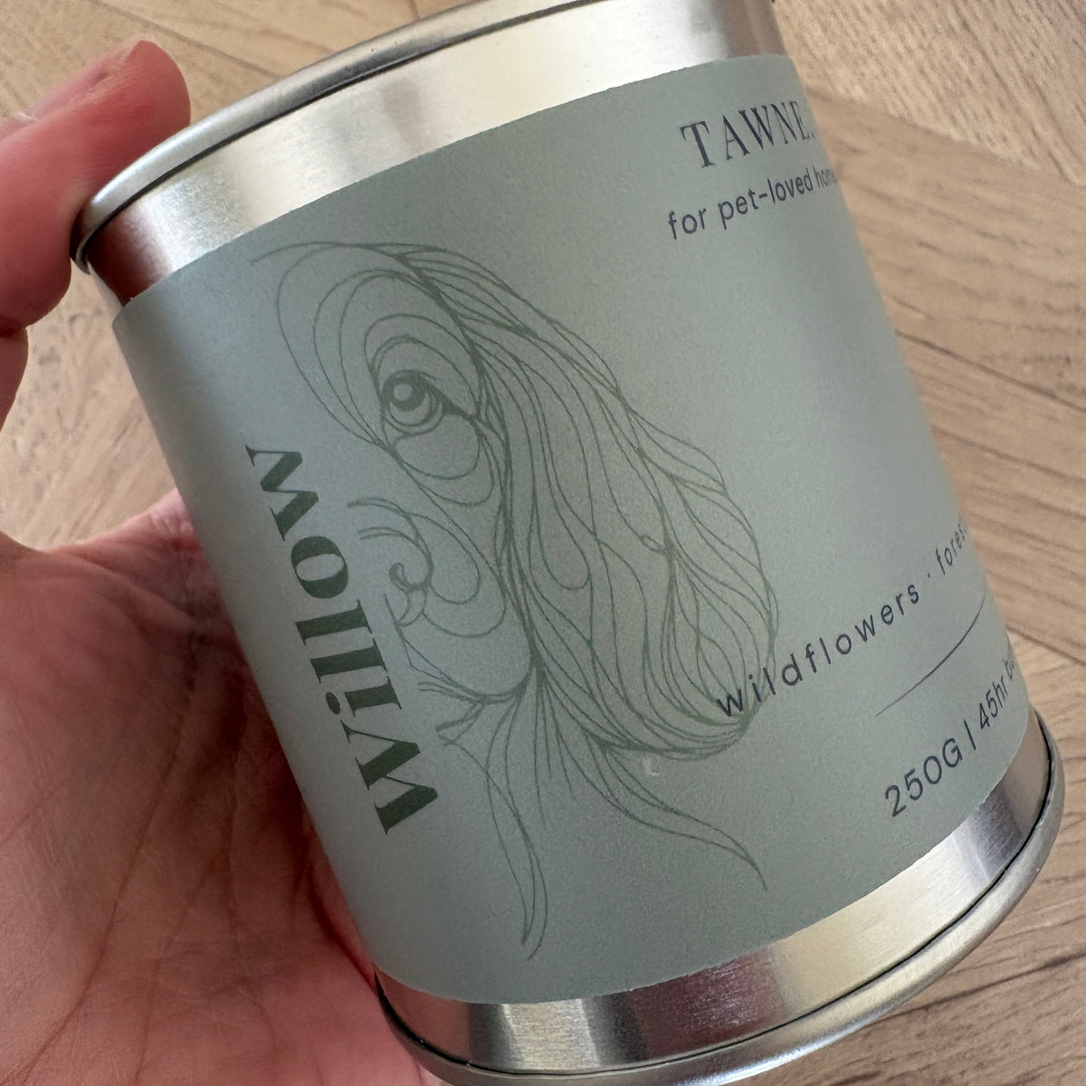 A hand holding a silver metal tin of Willow Tawne wildflowers pet candle
 against a light wood background.