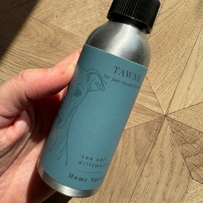 A hand holding a silver aluminum bottle of Tawne Sea Salt Driftwood Home Spray with a black cap and a dusty blue label, set against a wooden parquet floor.
