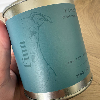 A hand holding a cylindrical metal tin candle, featuring a dusty teal label with a minimalist dog profile illustration.