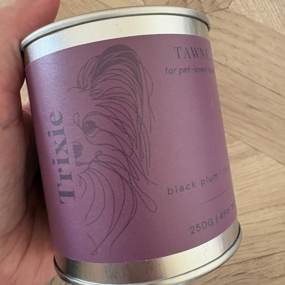 A hand holding a cylindrical metal can Trixie Tawne candle against a light wood parquet floor background.