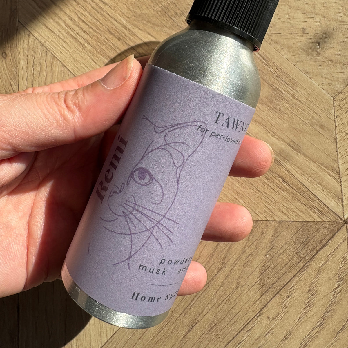A hand holding a silver spray bottle with a lavender label that reads 'TAWNE' and 'for pet-loved homes' with a minimalist cat line drawing. The background is a wooden floor.