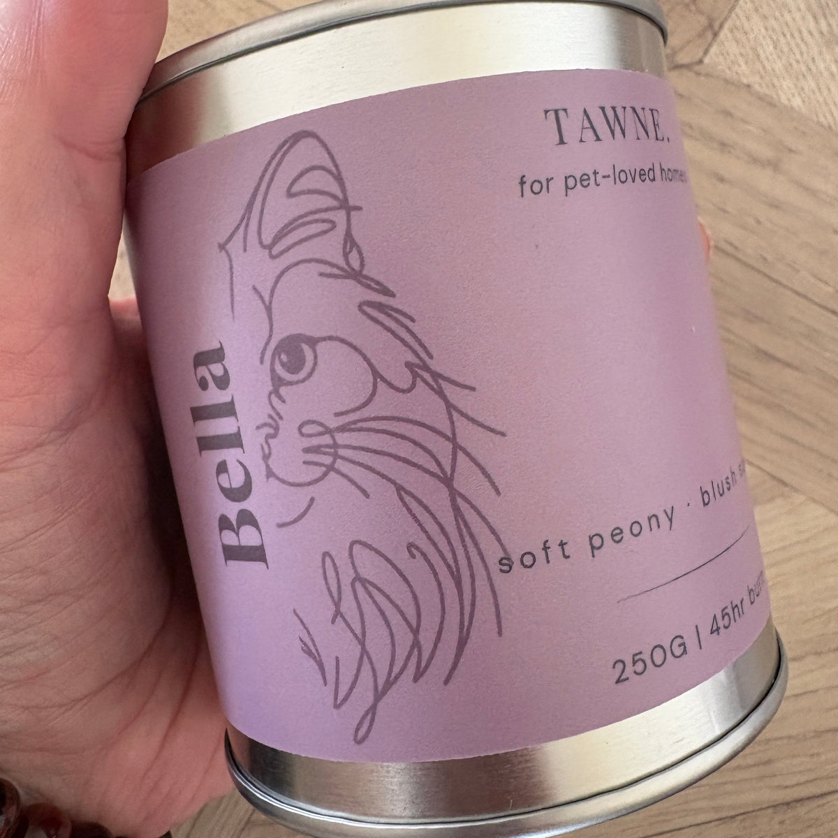 A hand holding a Bella Tawne candle. The can has a light purple label with a minimalist line art illustration of a cat and text reading 'Bella' and 'Tawne'.