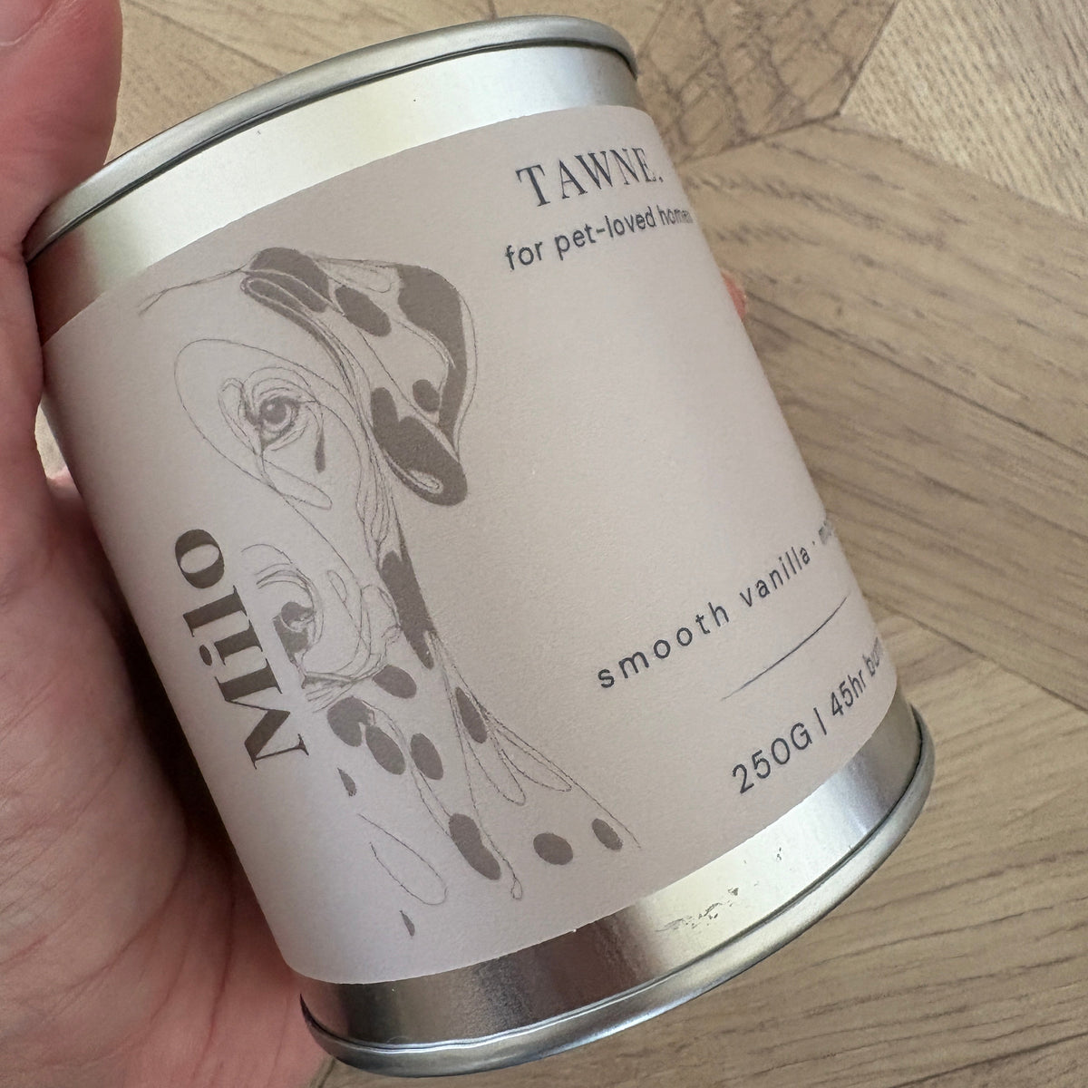 A hand holding a silver metal tin of Tawne smooth vanilla pet candle against a light wood parquet background.