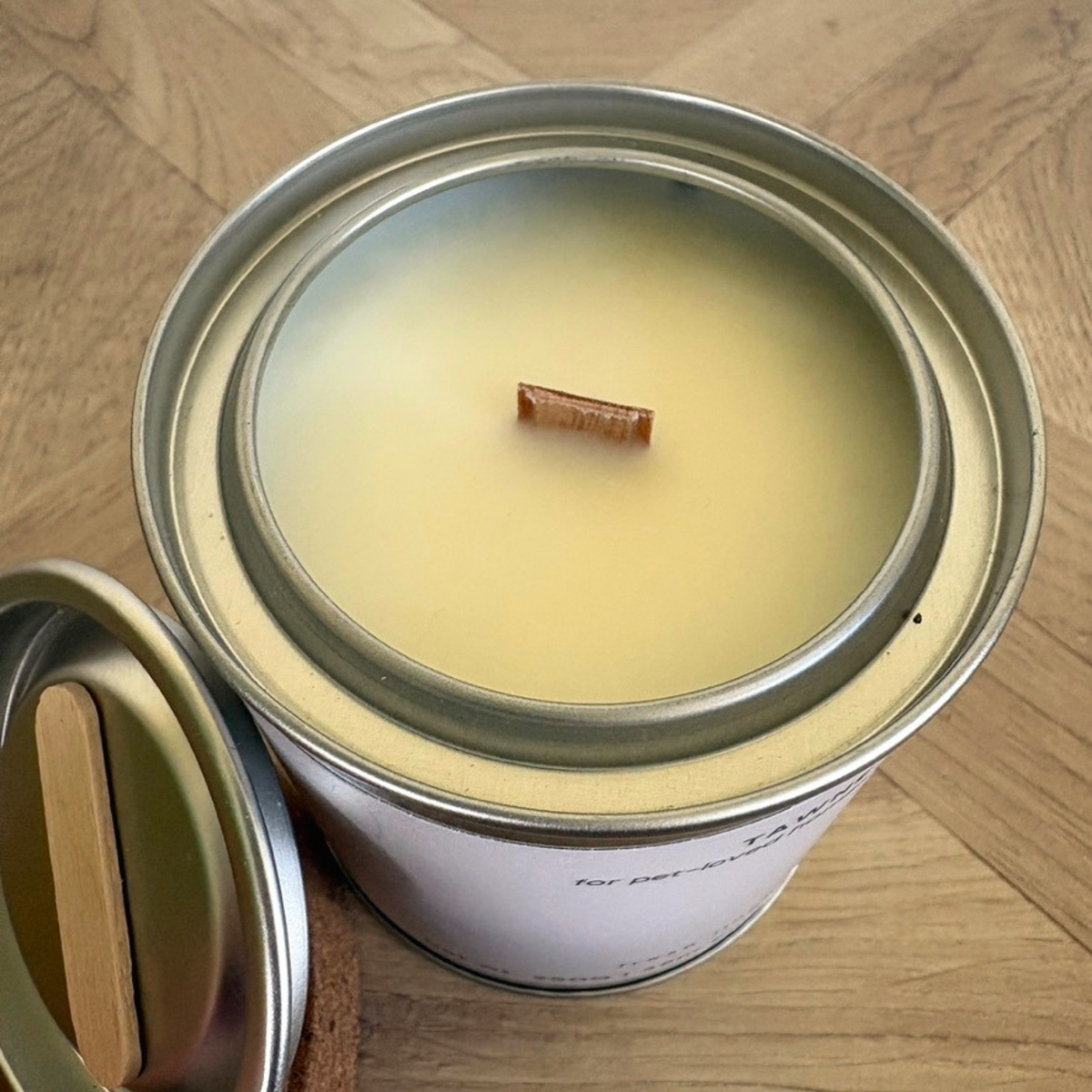 An open metal tin candle with pale creamy yellow wax and a small cinnamon stick wick, shown on a light wood floor with the lid placed beside it.