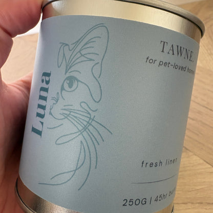 A hand holding a cylindrical metal tin candle. The tin has a pale blue label with a minimalist line drawing of a cat and the text 'Luna' and 'TAWNE for pet-loved homes fresh liner 250G'.
