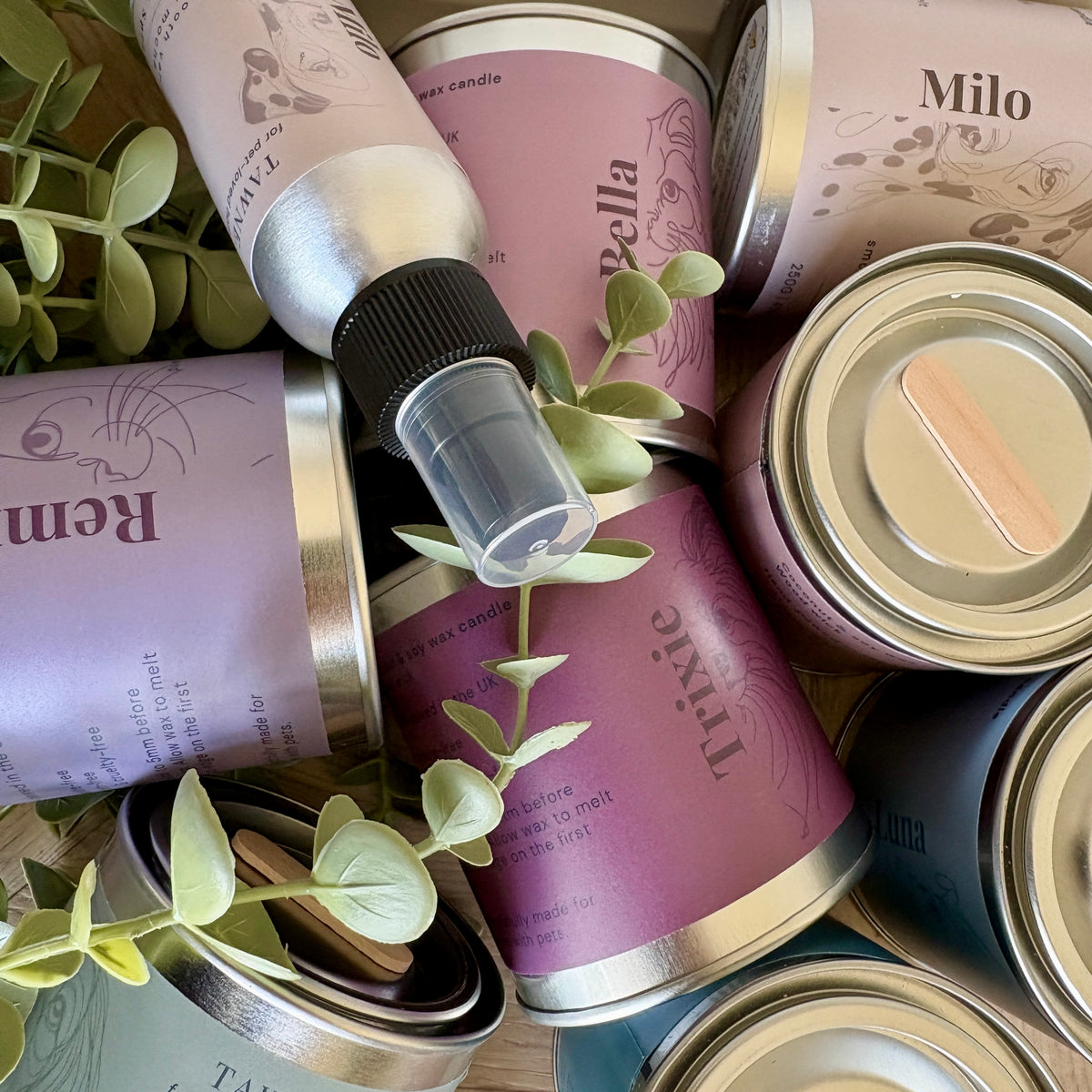 A styled flat-lay of multiple tawne candle tins and a spray bottle with lavender labels and silver metal cans, arranged with green eucalyptus leaves on a wooden surface. Visible text includes 'Willow & Bella', 'Remi', and 'Trixie', along with names like 'Milo'.