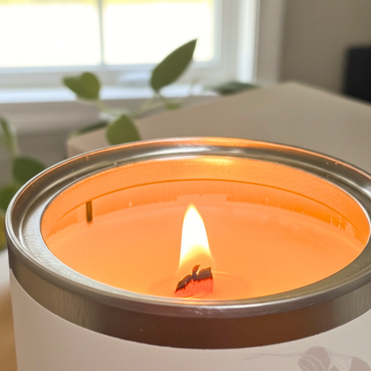 A close-up of a lit candle in a silver tin, showing a warm orange wax surface and a small flame, with a softly blurred indoor background.