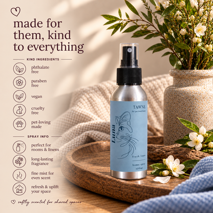 A silver spray bottle of Tawne Fresh Scents room spray with a light blue label, styled on a wooden tray with a potted plant and white blossoms in a warm, sunlit setting. Text on the left lists product benefits and ingredients.